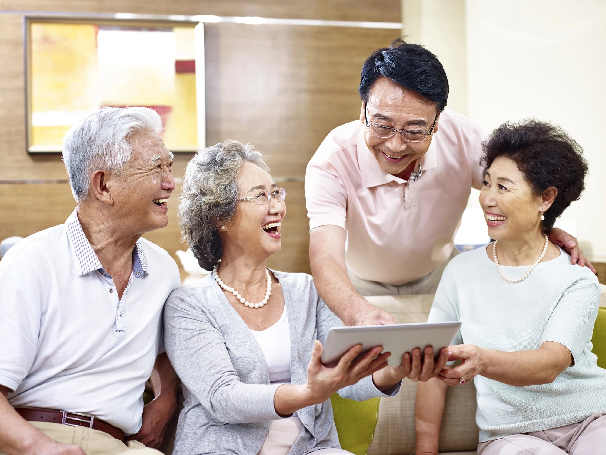 Group of happy Asian seniors laughing together while looking at a tablet in a bright living room