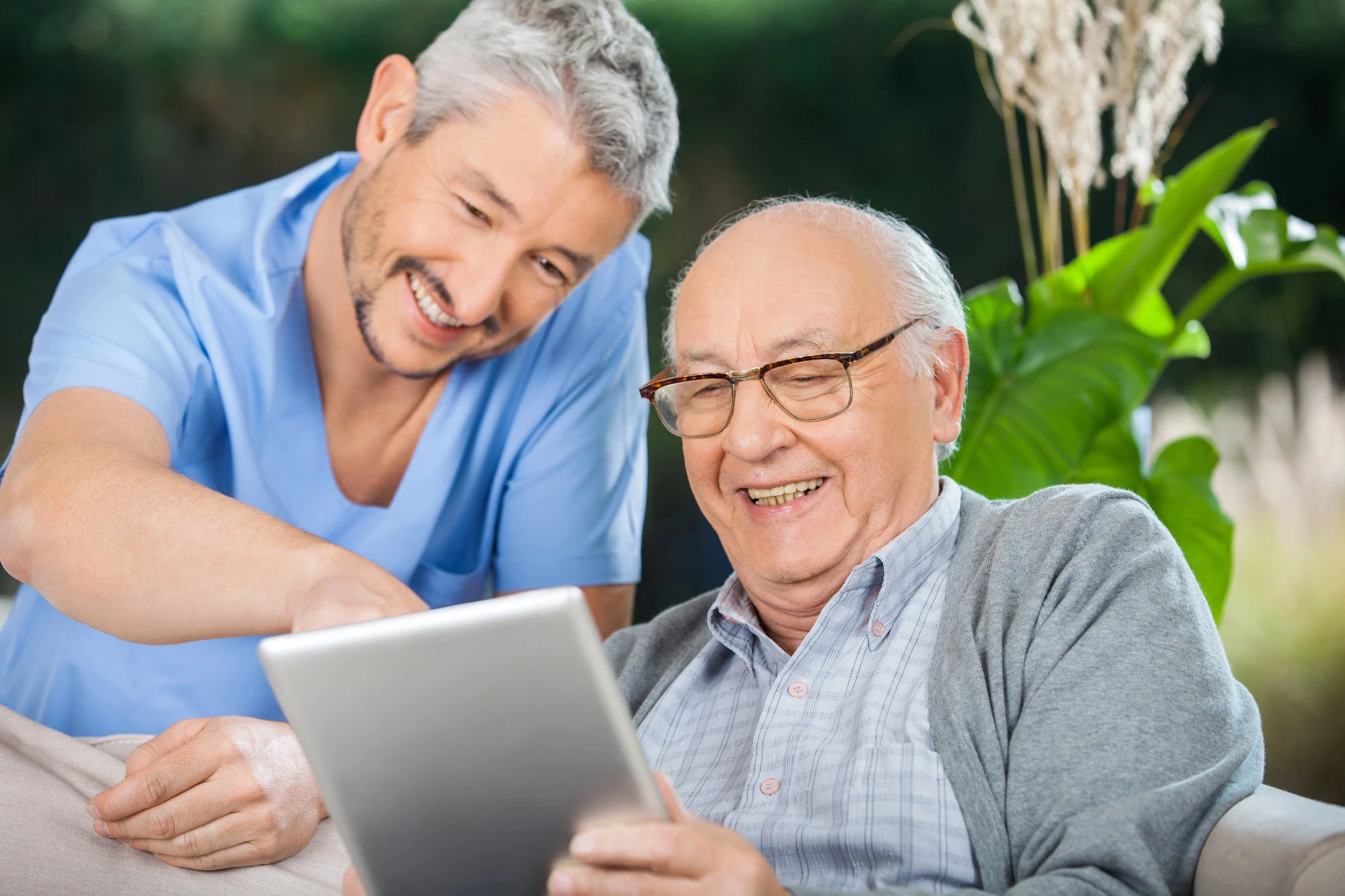 Smiling male caregiver in blue scrubs helping a happy elderly man use a tablet outdoors