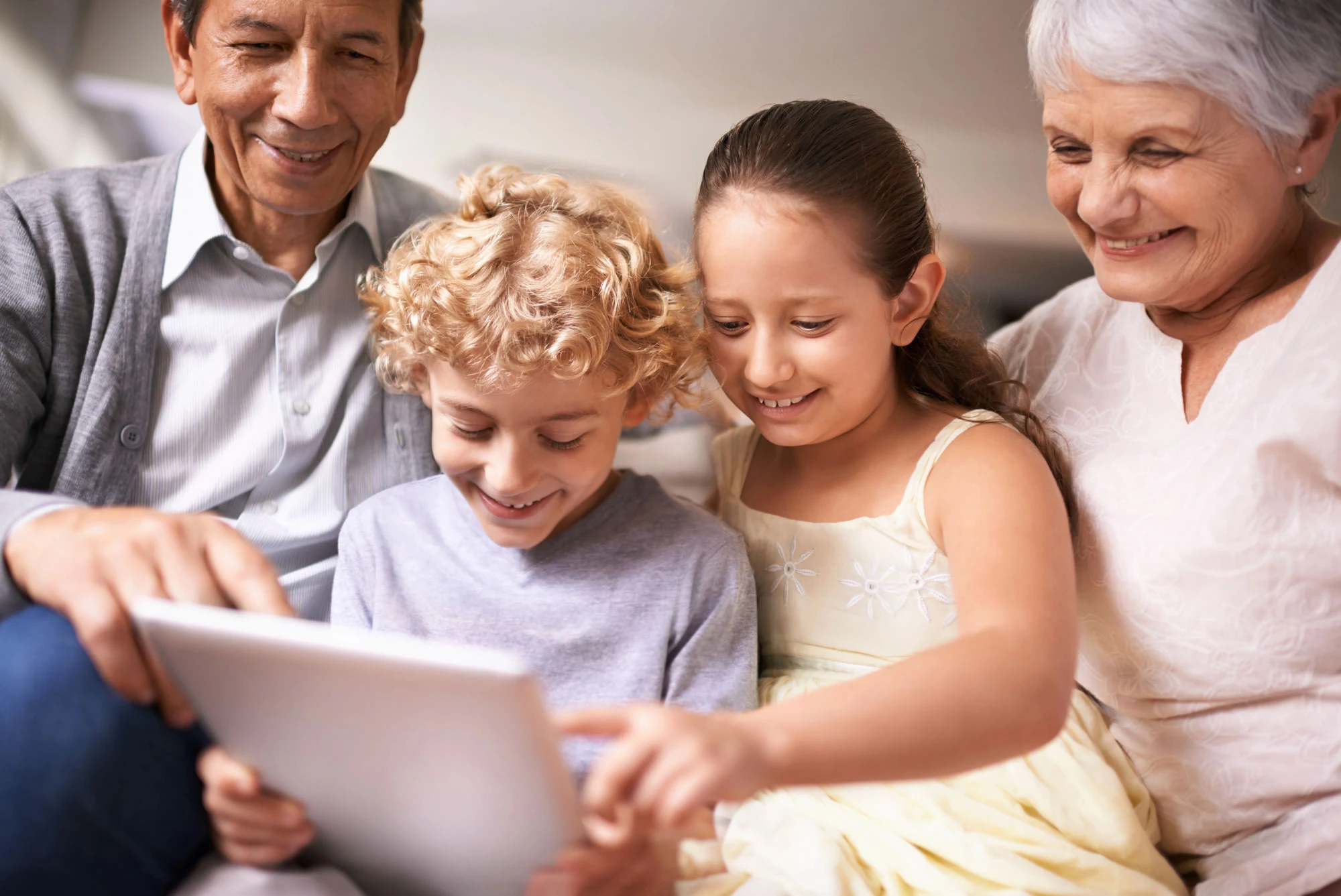 Happy multi-generational family: grandparents with young grandchildren smiling and using a tablet together on the couch